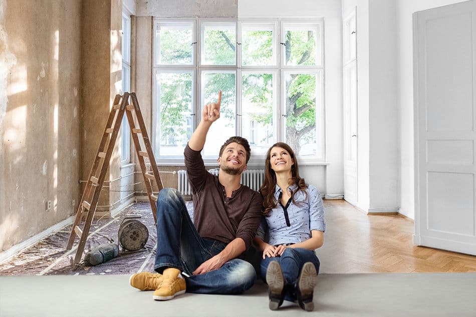 Een man en een vrouw zitten op de vloer van hun woning die ze aan het verbouwen zijn. De man lacht en wijst naar boven.